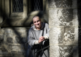 Photo of Andrea in front of the stone walls of a church. She has a shaved head and is smiling at the camera, sitting on a step