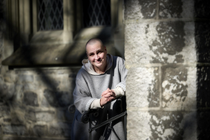 Photo of Andrea in front of the stone walls of a church. She has a shaved head and is smiling at the camera, sitting on a step