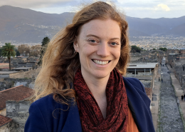 Photo of Robyn Thomas smiling with hills and houses behind her. She has long auburn hair and is wearing a blue jacket with a red scarf around her neck and an orange top