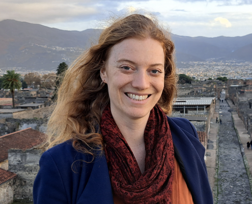 Photo of Robyn Thomas smiling with hills and houses behind her. She has long auburn hair and is wearing a blue jacket with a red scarf around her neck and an orange top