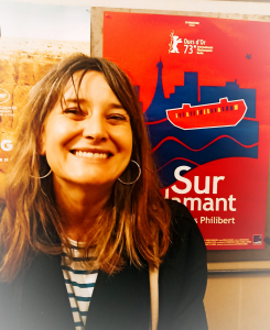 Photo of Liza smiling with a breton top, long brown hair and standing in front of a poster of the film Sur L'Adamant