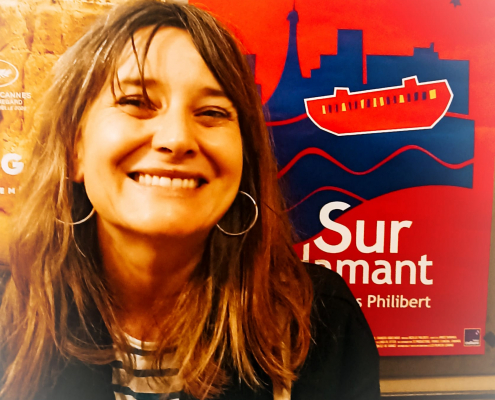 Photo of Liza smiling with a breton top, long brown hair and standing in front of a poster of the film Sur L'Adamant