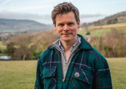 Will with the Welsh hills behind him wearing a check jacket and open necked shirt