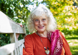 Emmy sitting on a bench with green trees behind her, looking at the camera with twinkling eyes and silver hair wearing her trademark red with long necklaces around her neck